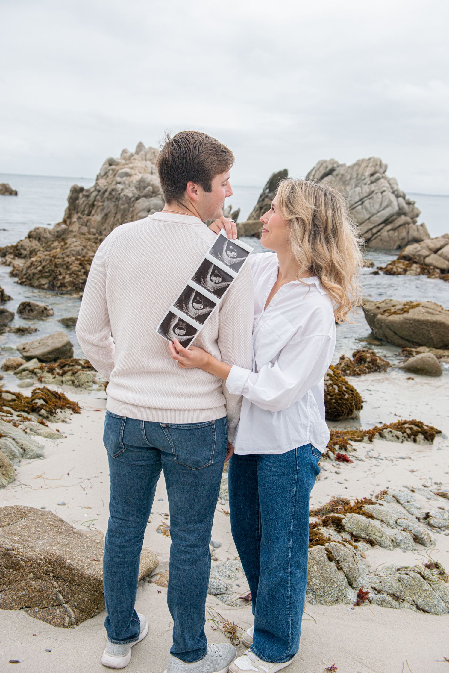 Couple sharing a tender moment on a rocky beach, holding ultrasound photos, announcing their pregnancy.