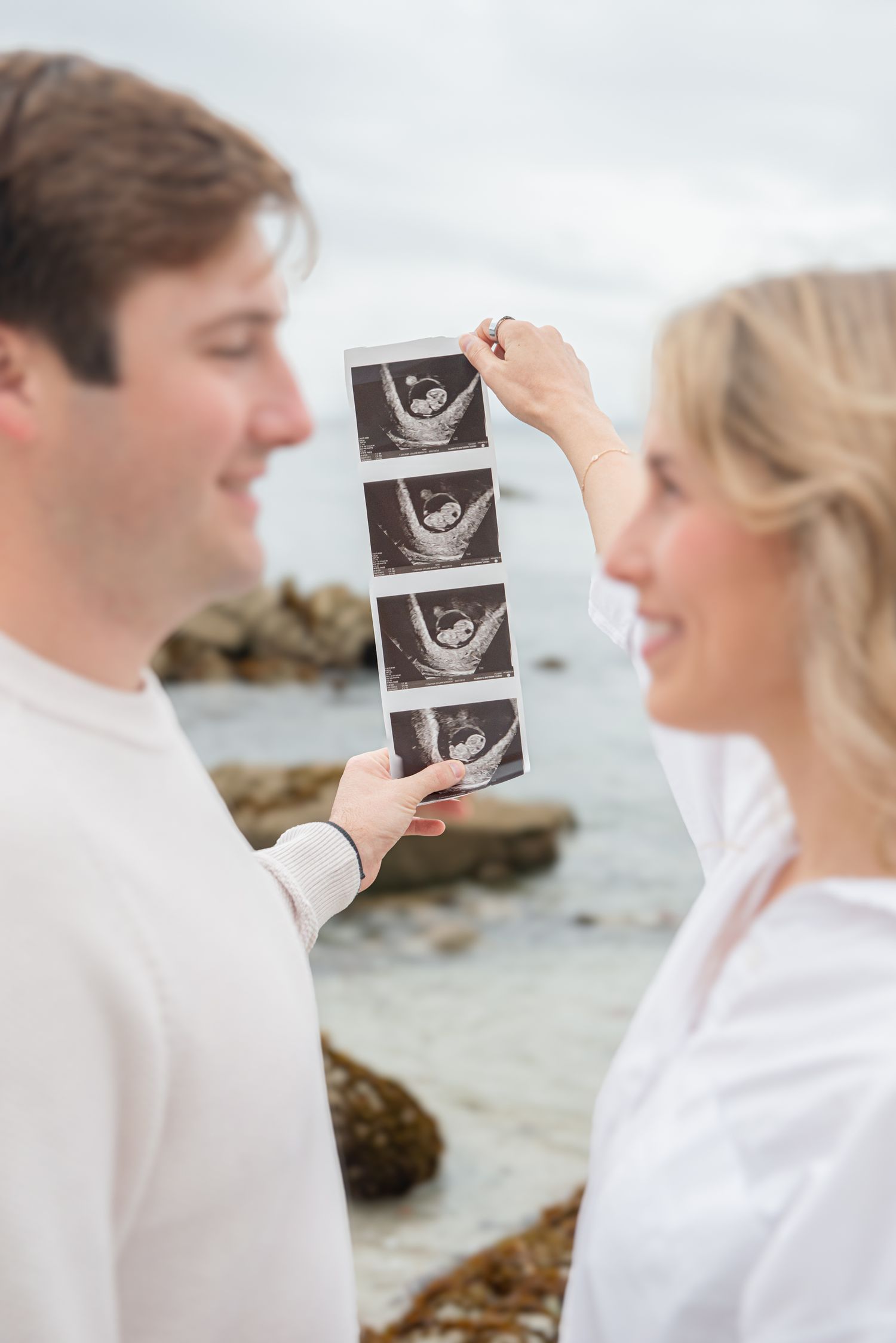 Smiling couple holding ultrasound photos by rocky beach, celebrating pregnancy announcement outdoors.
