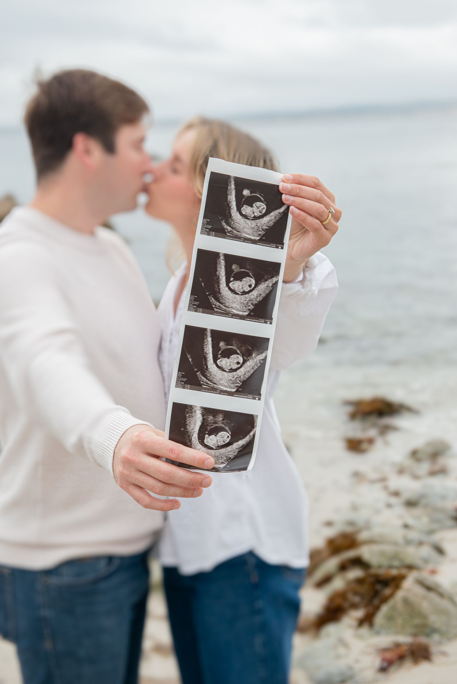Couple kissing on rocky beach holding ultrasound photos, announcing pregnancy with ocean backdrop.