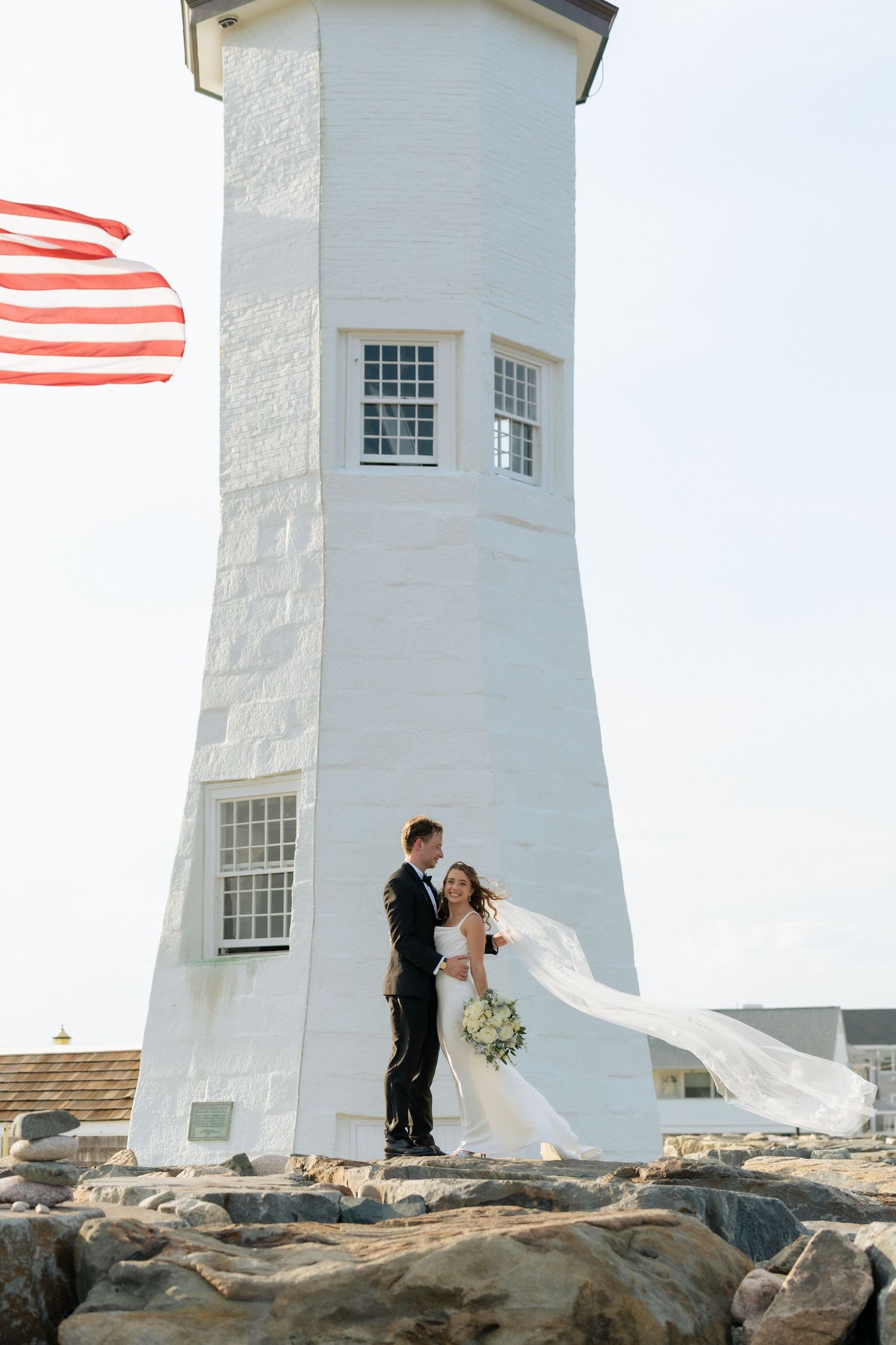 Bride and groom embrace at base of white lighthouse with American flag, bridal veil flowing in wind on rocky New England coast.