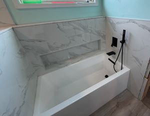 White marble tiled bathtub area with built-in shelf niche and matte black handheld shower fixture.