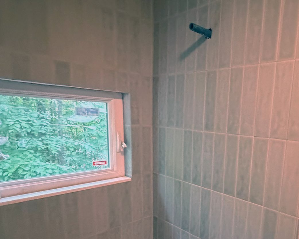 Tiled bathroom wall with a small window overlooking greenery and a metal shower fixture installed.