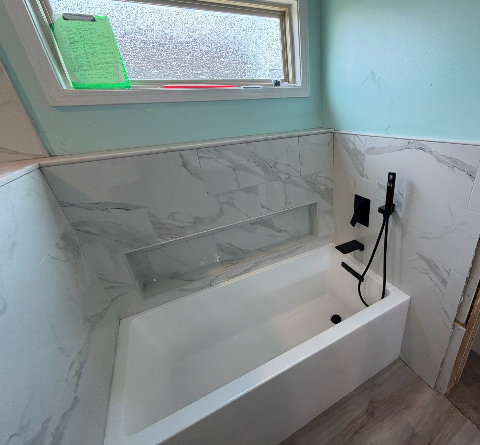 Modern white marble tiled bathtub surround with matte black faucet and small window.
