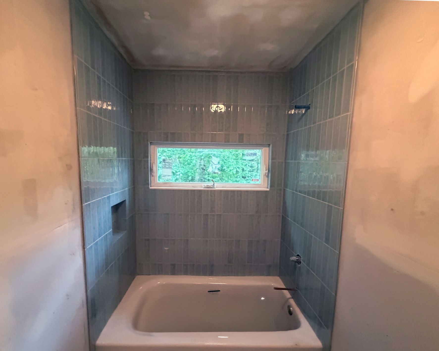 Partially completed blue-gray tiled bathtub surround with small window during bathroom renovation.
