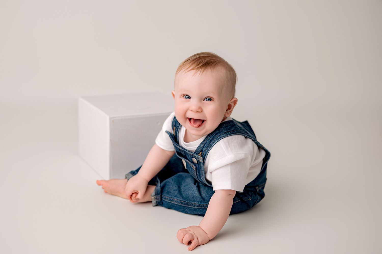 Smiling baby girl in white lace dress and floral crown sitting in a rattan basket on a rustic wooden floor.