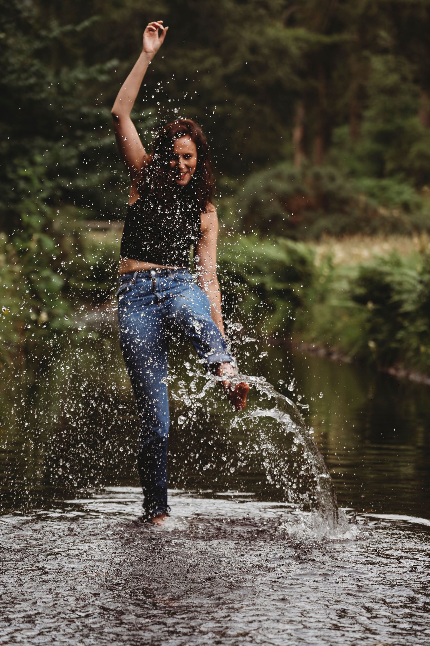 A joyful young woman splashing water in a river, wearing jeans and a black top, surrounded by lush green forest.