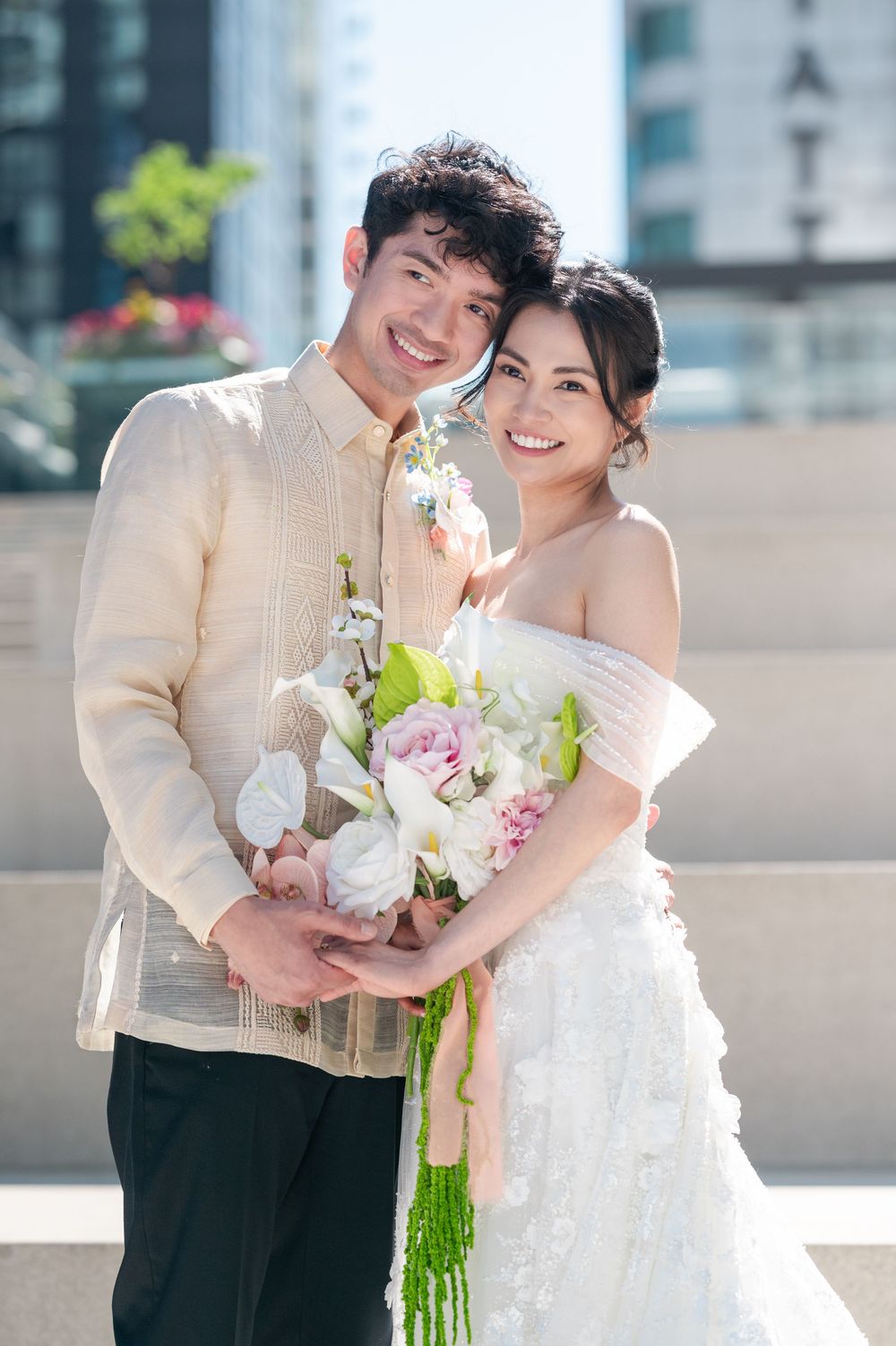 A smiling couple in wedding attire holds a bouquet of pink and white flowers outdoors with city buildings in the background.