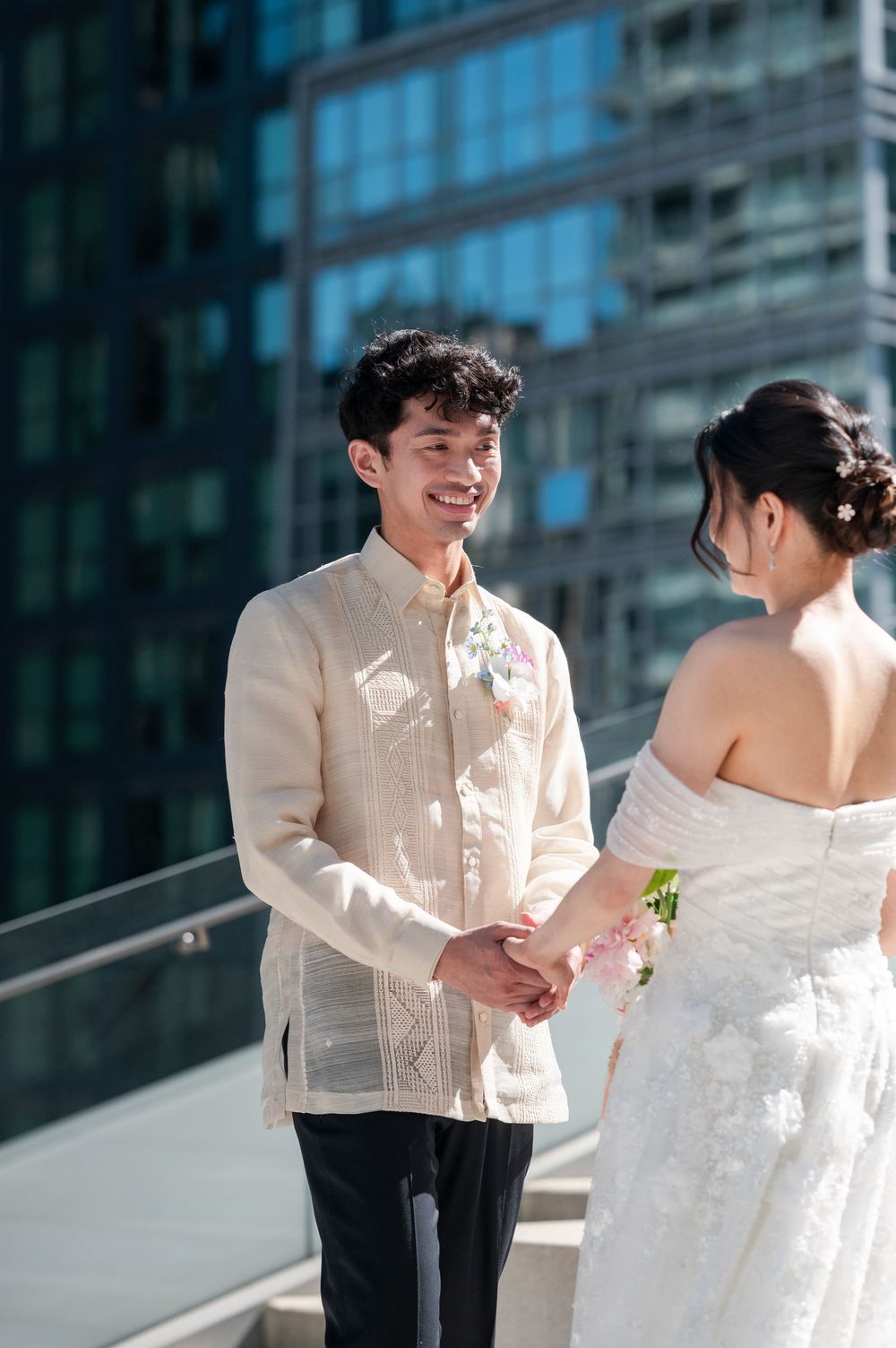 Smiling groom in barong tagalog holds hands with bride in white off-shoulder gown on urban rooftop.