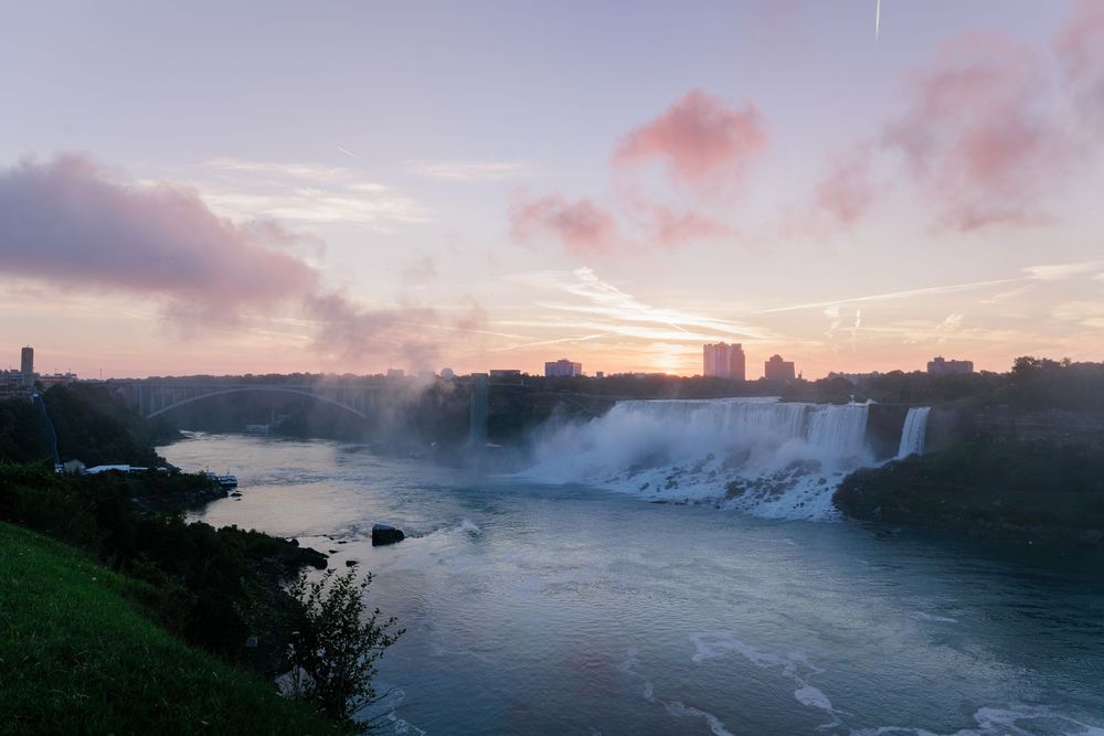 Aerial view of Niagara Falls at sunset with mist rising over turquoise waters and city skyline in the background.