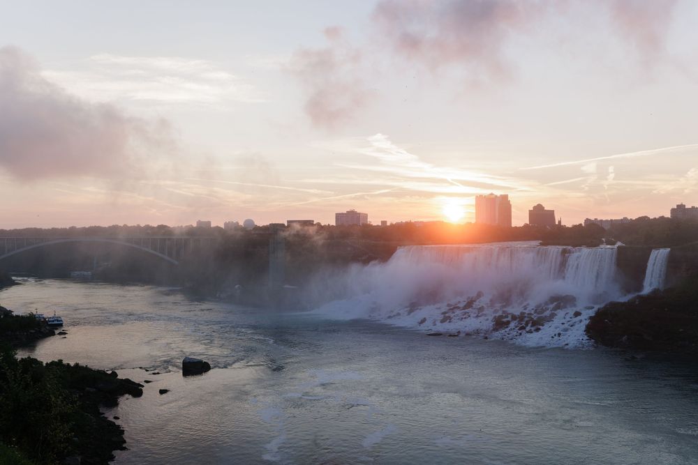 Stunning sunrise over Niagara Falls with mist rising from cascading waterfalls and city skyline in the background.