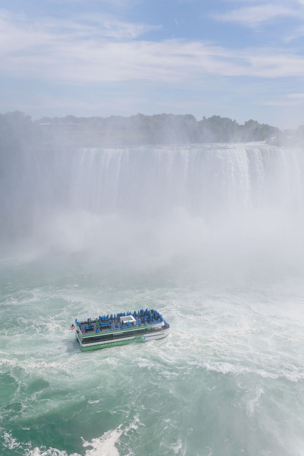 Tourist boat navigates misty turquoise waters near the base of massive Niagara Falls on a partly cloudy day.