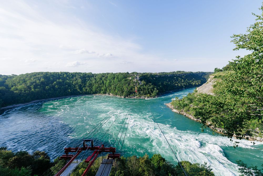 Aerial view of the Niagara River rapids with turquoise rushing water, lush green trees, and a red structure on a sunny day.