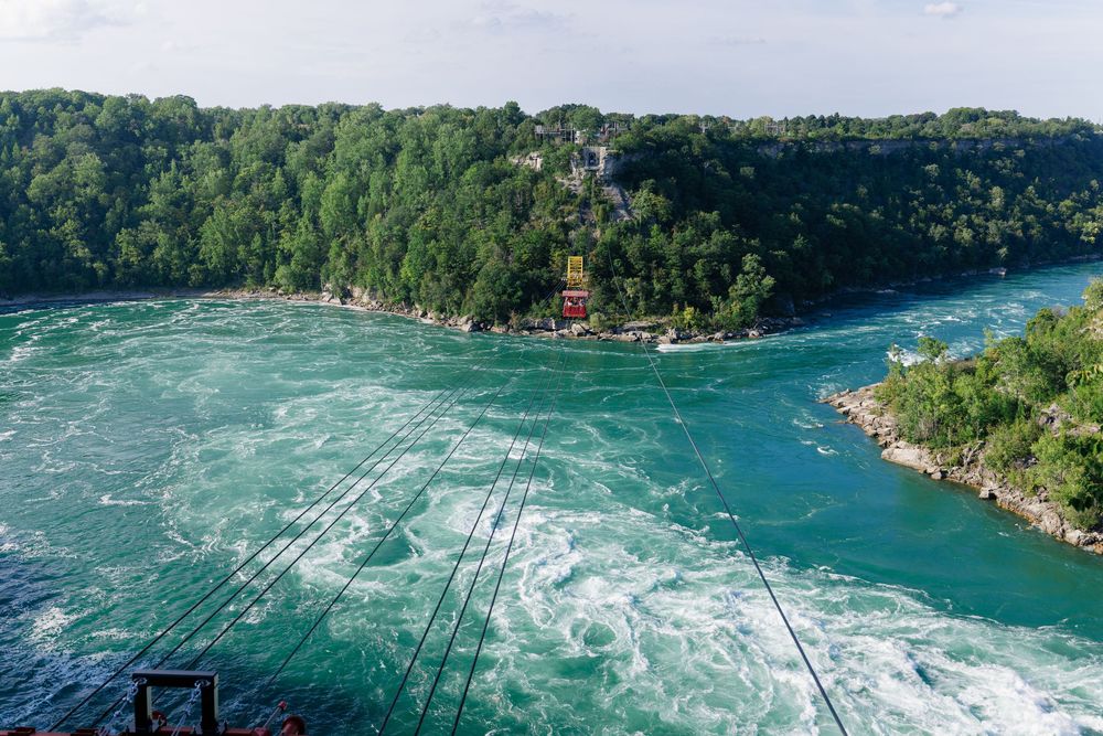 Aerial view of turquoise Niagara River rapids with lush green forest and a red building on the rocky shoreline.