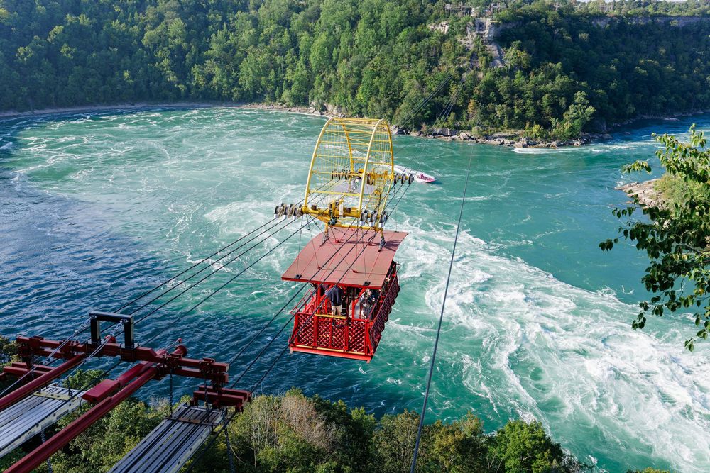 Red cable car gondola crossing turquoise Niagara Whirlpool Rapids on the Whirlpool Aero Car, surrounded by lush green forest.