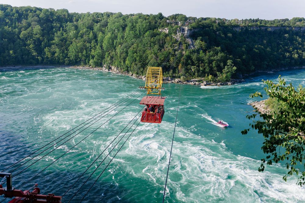 Red cable car gondola crossing turbulent Niagara River gorge with lush green cliffs and a boat below.