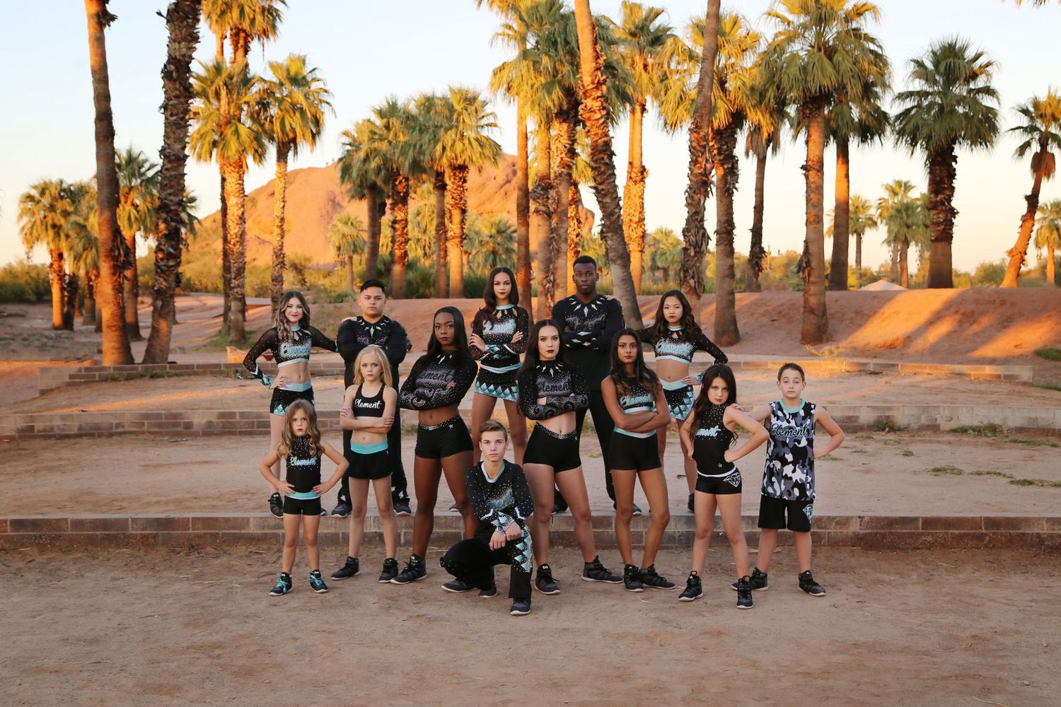 A group of young cheerleaders in black athletic uniforms pose together among tall palm trees with a desert mountain behind them.