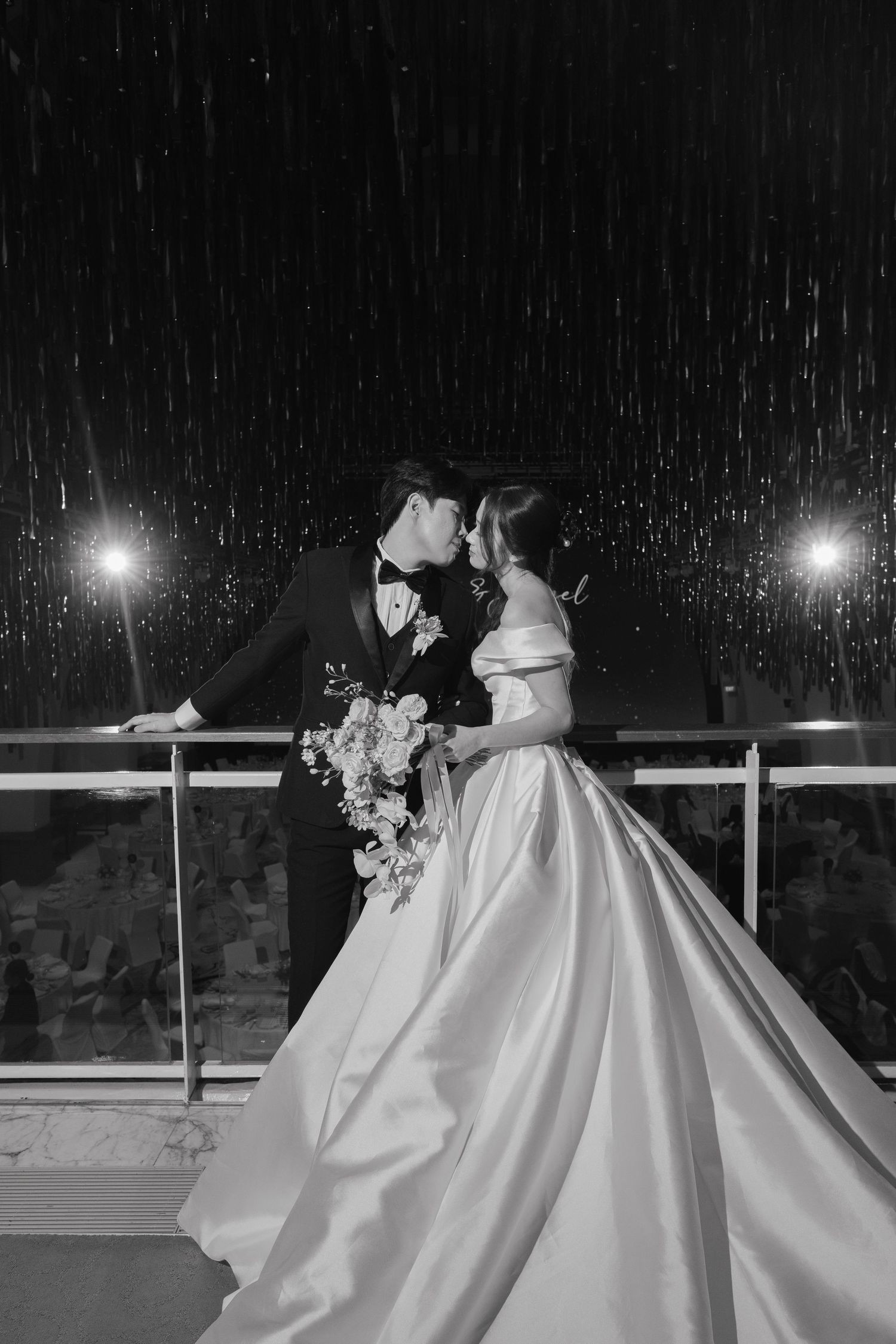 Black and white photo of a bride and groom sharing a romantic kiss in the rain at night, bride holding a floral bouquet.