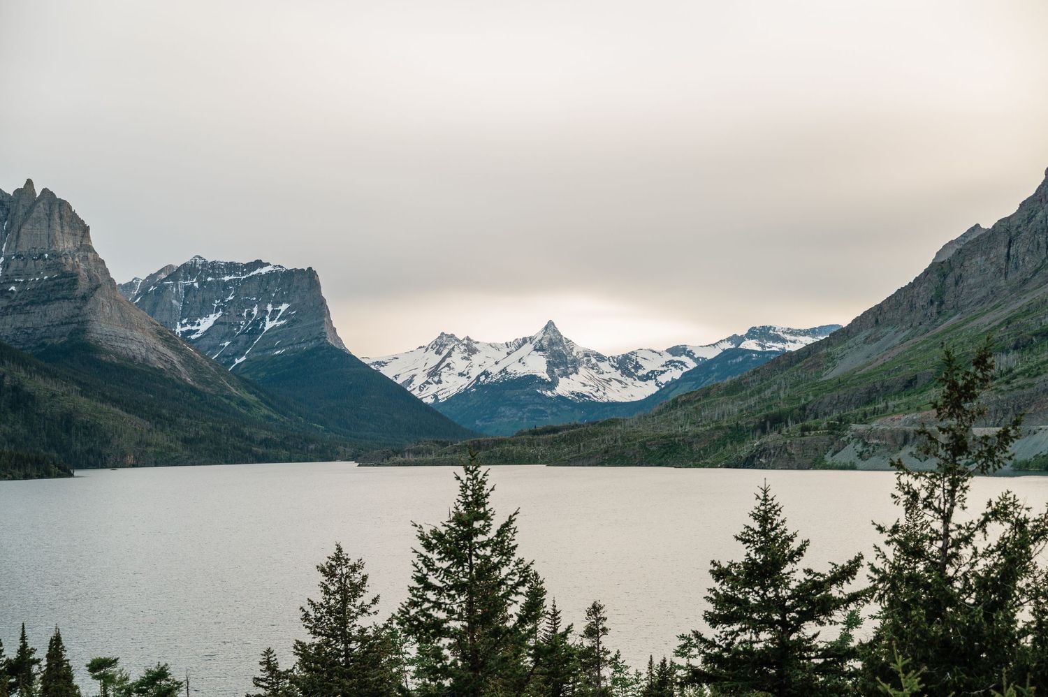 Snow-capped mountains reflect over a calm alpine lake surrounded by evergreen trees under overcast skies.