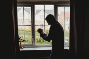 Silhouette of a person holding a baby while standing by a window with houses visible outside.