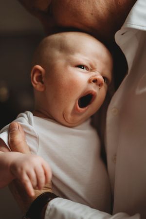 A tiny baby yawning while wearing a white onesie in a close-up portrait shot.