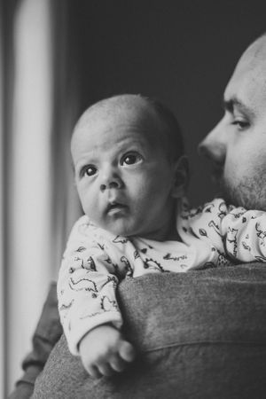 Black and white portrait of a wide-eyed infant being held against a shoulder near a window.