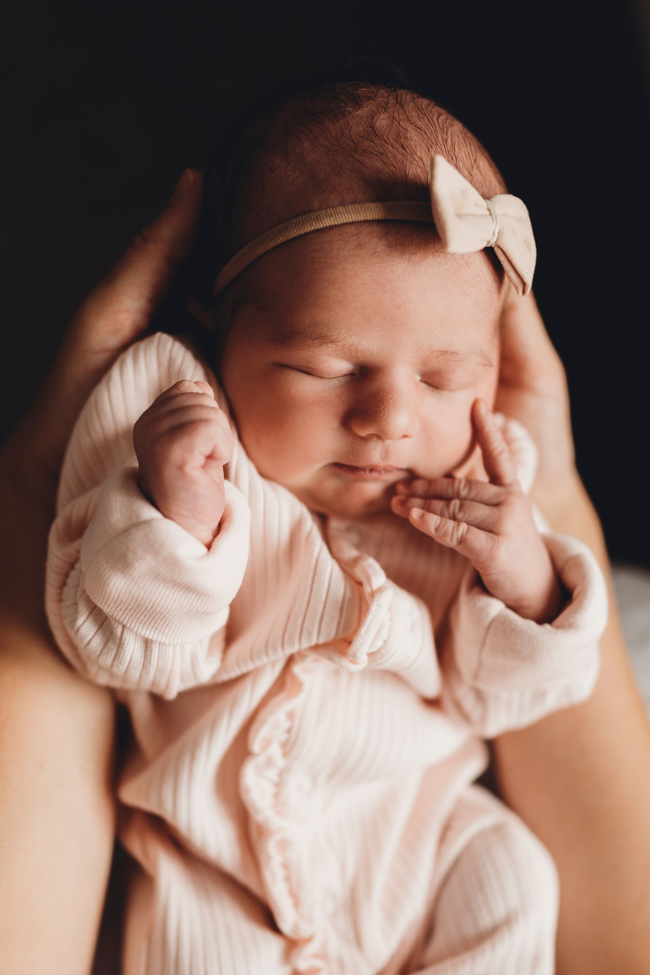 Sleeping newborn baby wearing pink with a delicate bow headband against dark background.