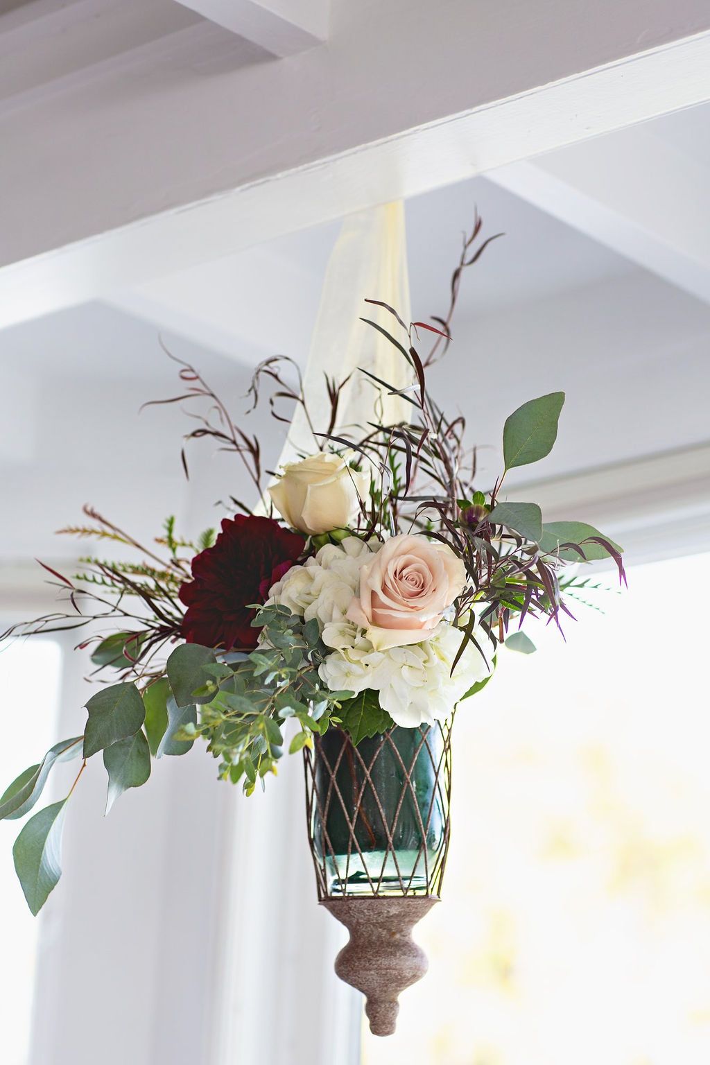 Bride in white gown and bridesmaid in grey dress sitting together holding white floral bouquets indoors.