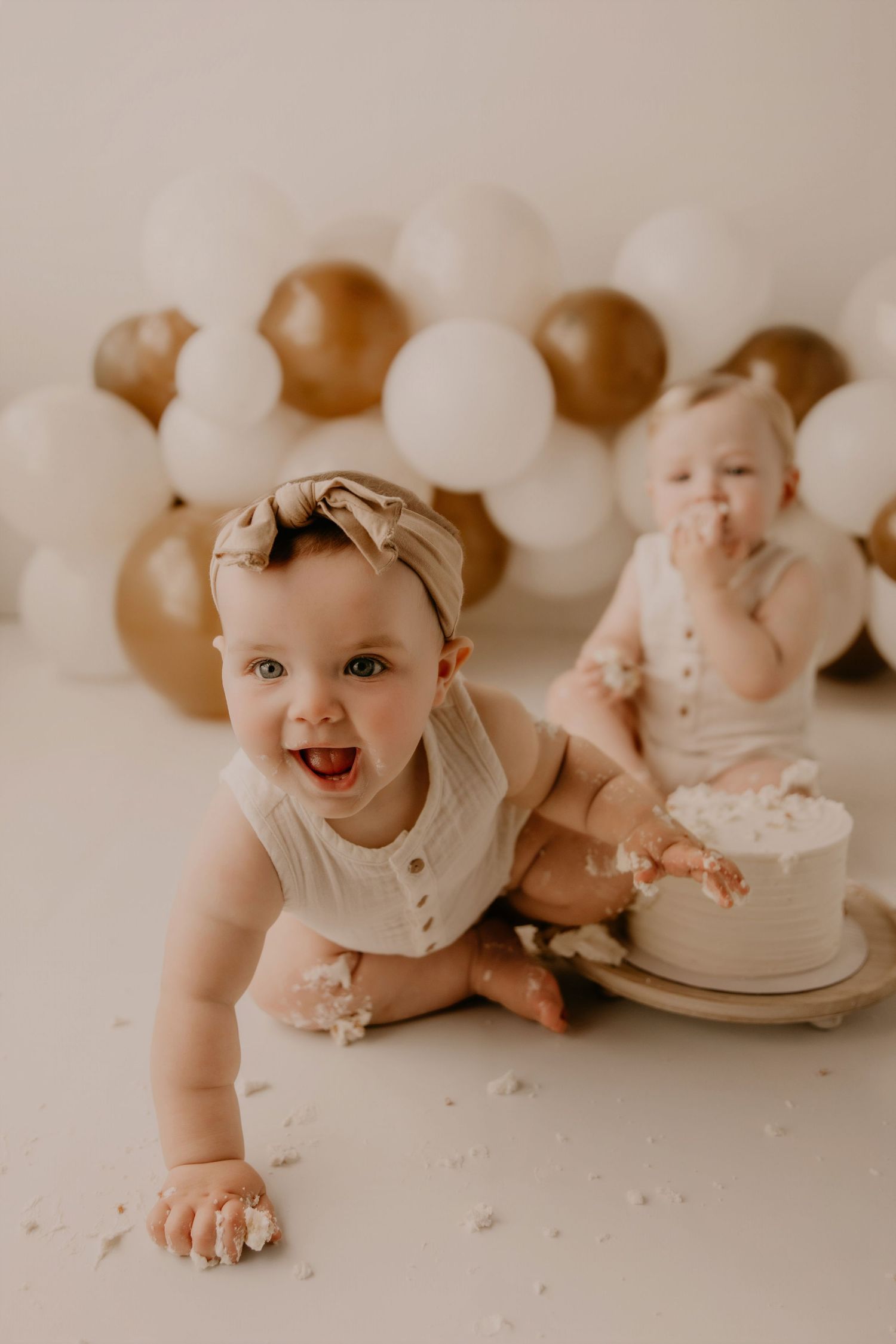 Two babies in cream rompers enjoy a cake smash birthday photo session with gold and white balloons backdrop.