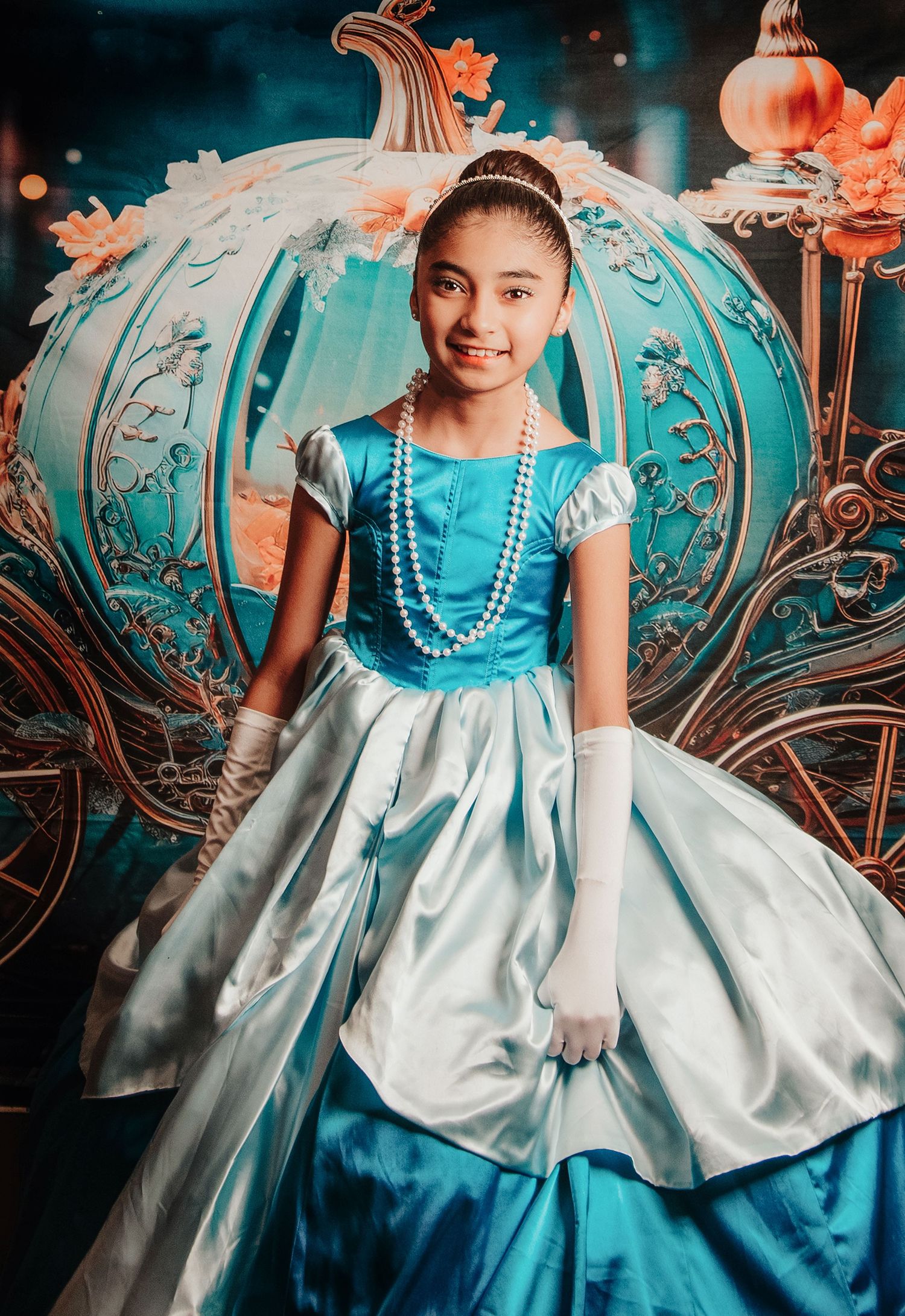 Young girl in Cinderella blue princess gown with pearl necklace and white gloves poses before a teal pumpkin carriage backdrop.