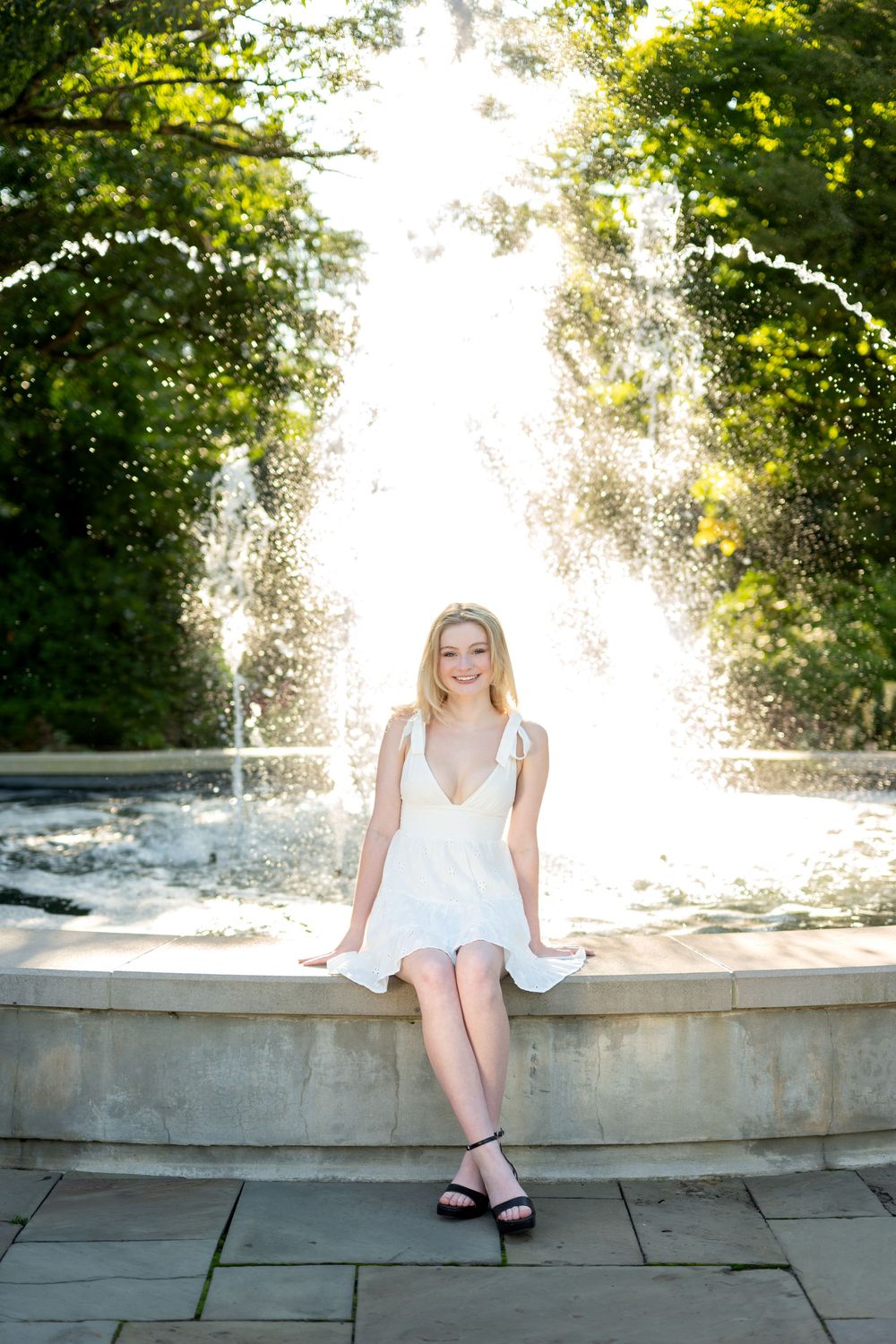 Smiling blonde woman in white dress sitting on fountain edge in sunny park.