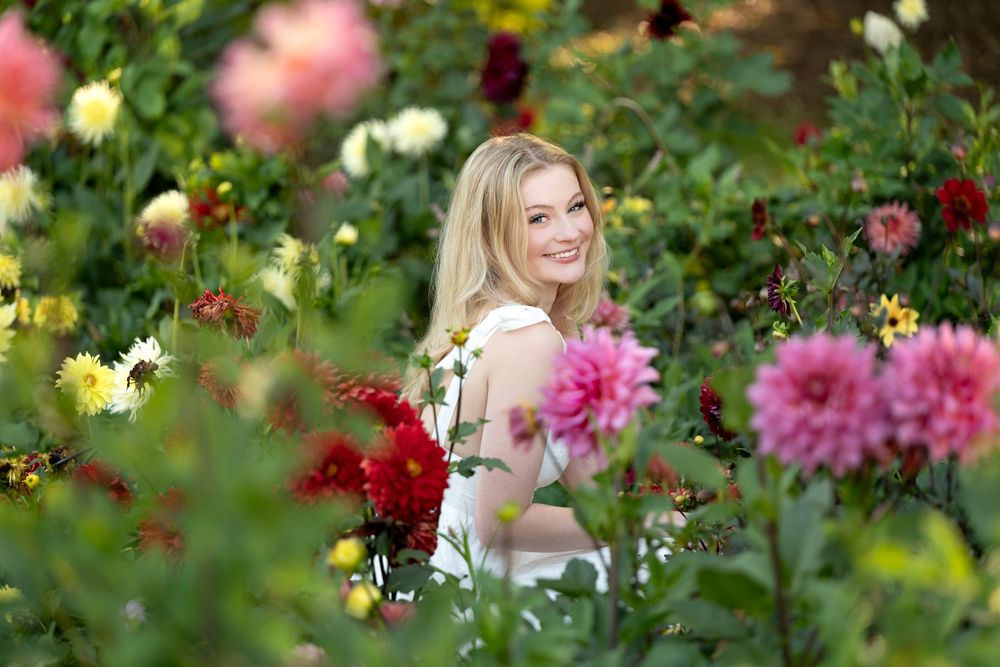 Smiling blonde woman surrounded by colorful dahlia flowers in garden.