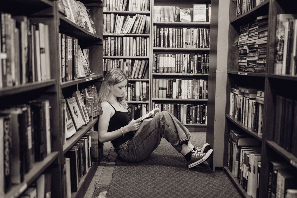 A young woman sits on a bookstore floor reading a book surrounded by tall shelves of books in black and white.
