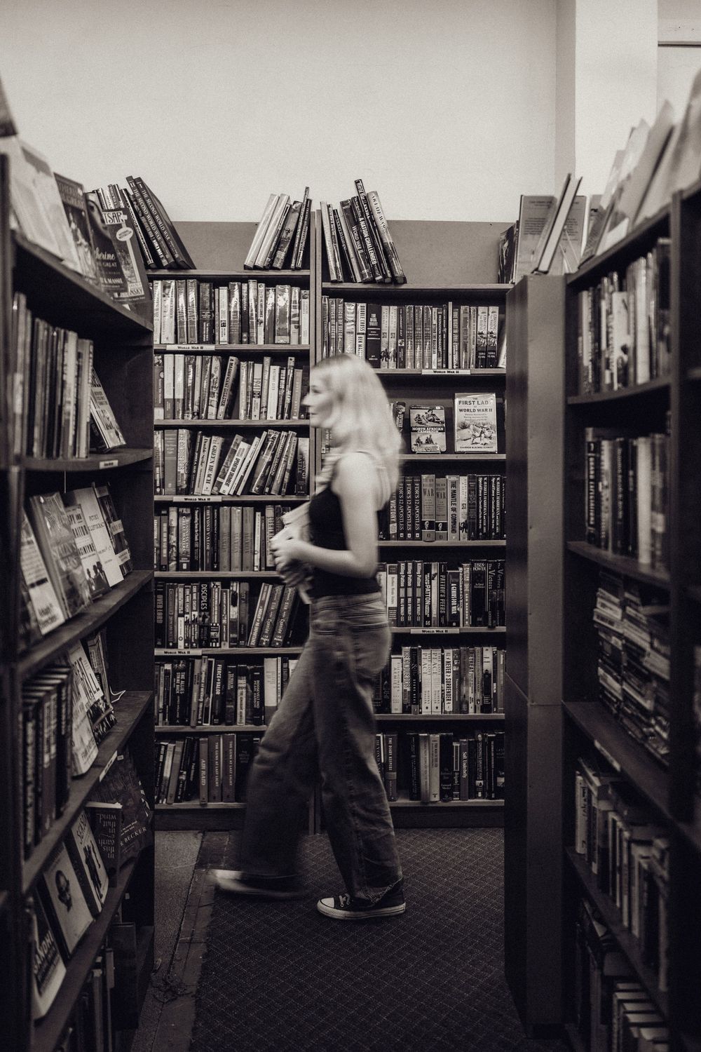 Black and white photo of a blonde woman browsing books in a library aisle lined with full shelves.