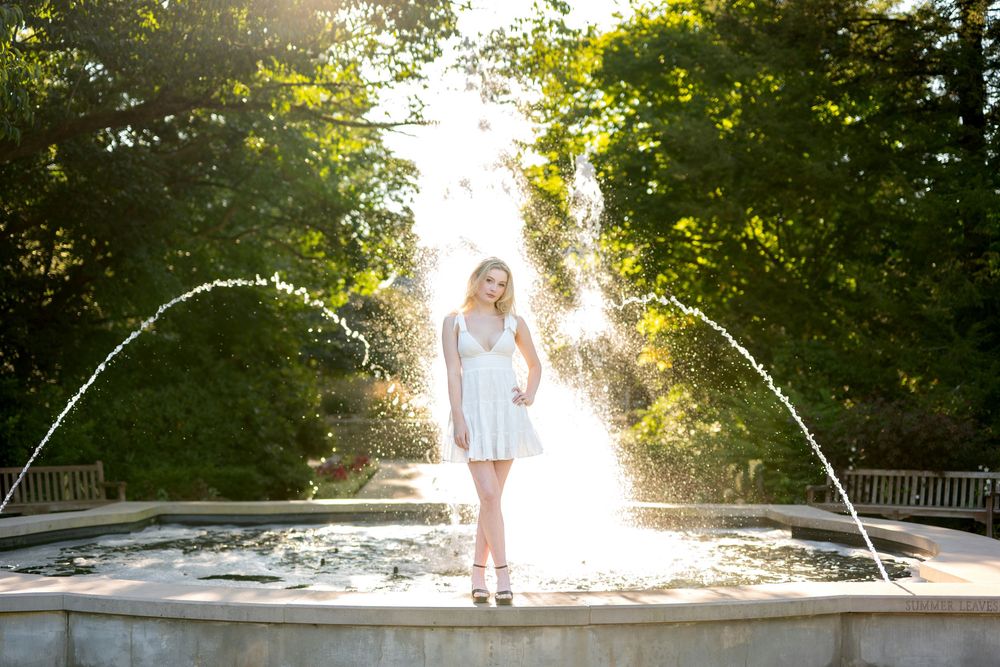 Blonde woman in white dress standing by fountain in sunlit park.