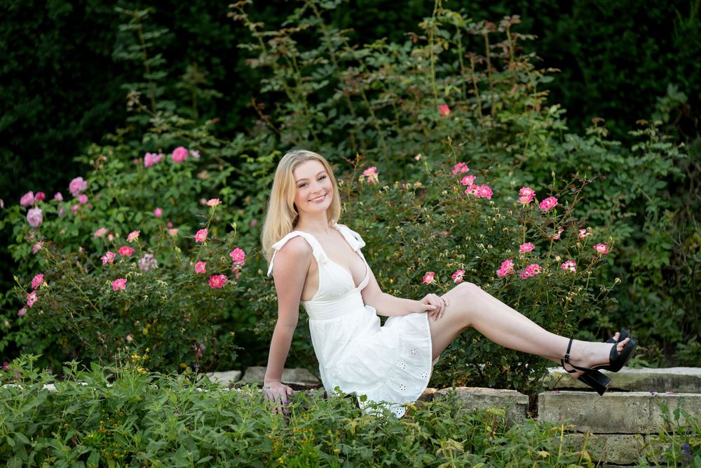 Young blonde woman in white dress sitting on stone wall surrounded by pink roses and lush green garden.