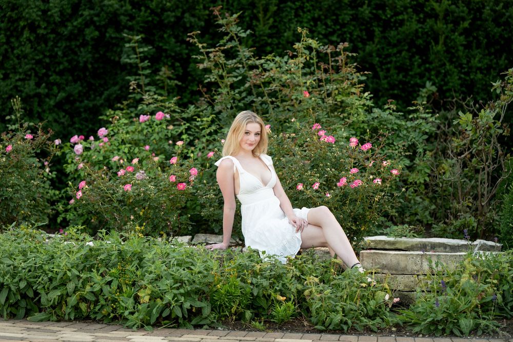 Blonde woman in white dress sitting on stone ledge surrounded by lush green shrubs and pink roses.