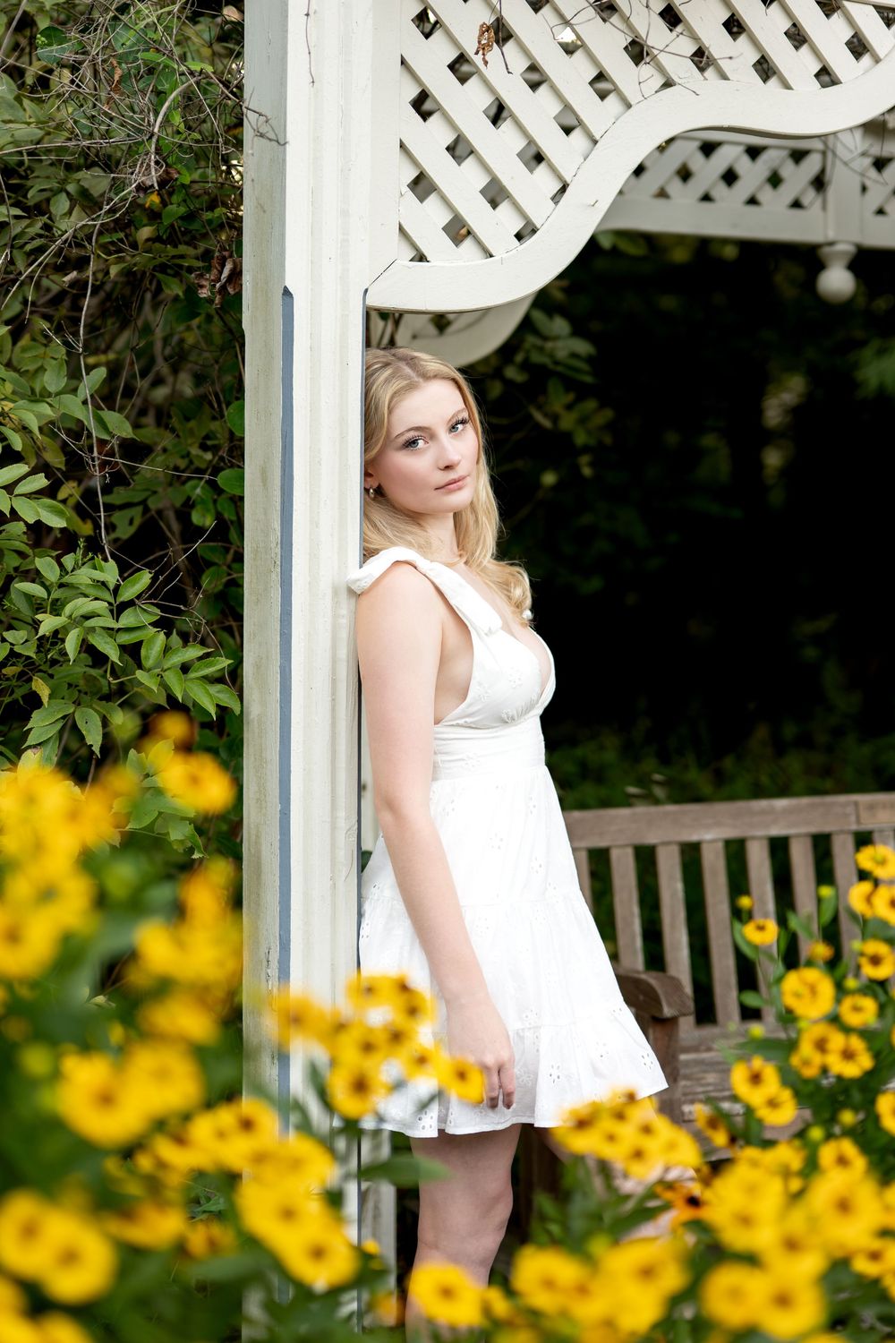 Blonde woman in white dress leaning against gazebo pillar surrounded by yellow flowers in a garden.
