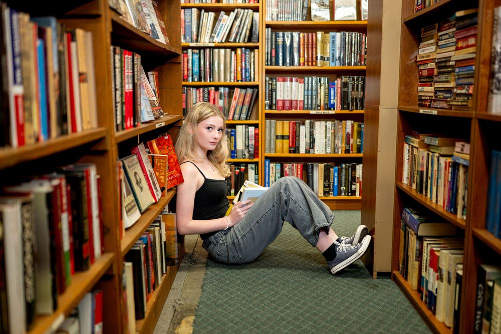 Young woman sitting on floor between bookshelves in a cozy library, holding books, wearing jeans and sneakers.