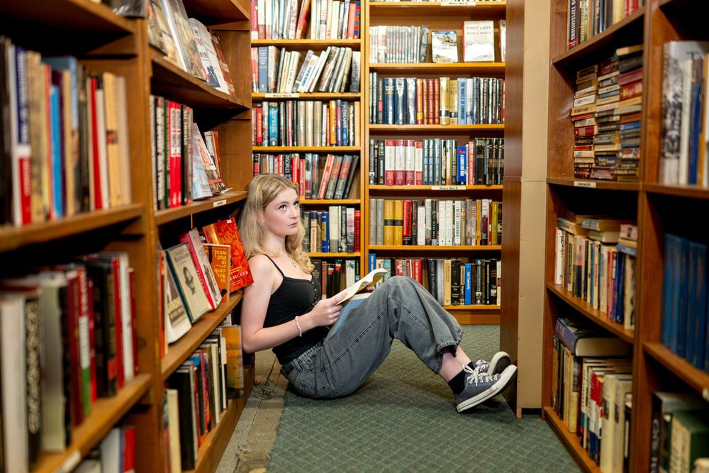 Young blonde woman sitting on floor between tall wooden bookshelves in a cozy library, reading a book.