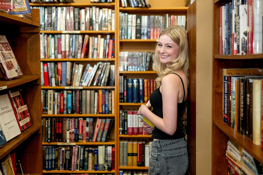 Young blonde woman smiling among tall wooden bookshelves filled with books in a cozy bookstore.