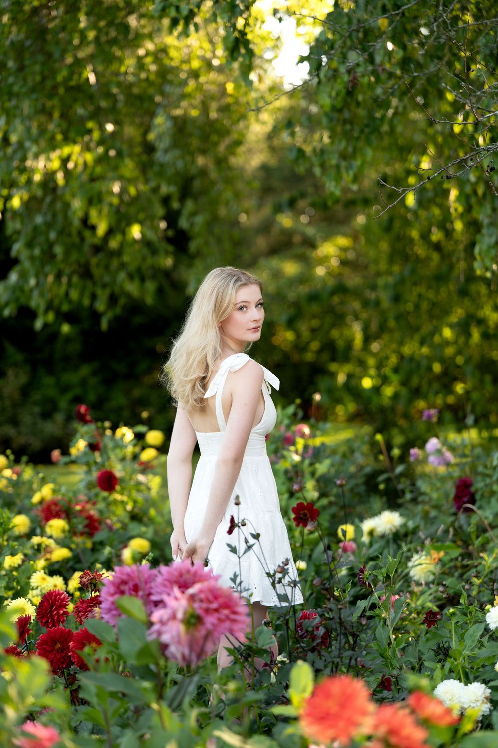 Blonde woman in white dress standing among colorful dahlias in a lush garden.