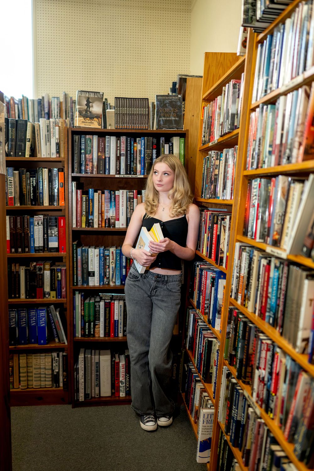 Blonde woman holding books while browsing shelves in a cozy, well-stocked bookstore aisle.
