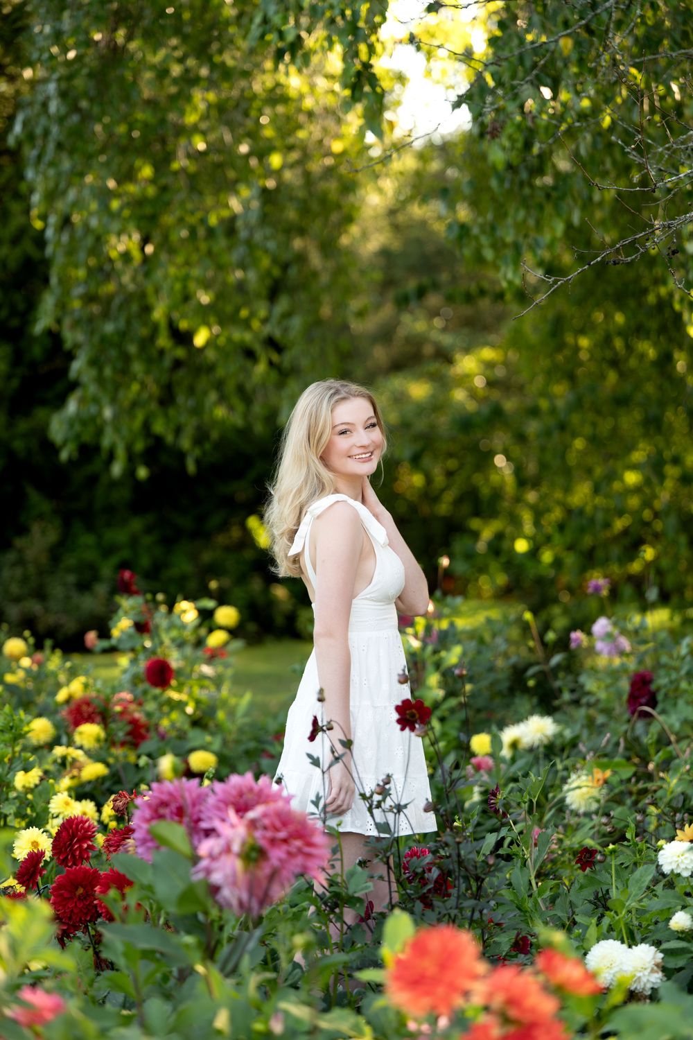 Smiling blonde woman in white dress stands among colorful dahlia flowers in sunny garden.
