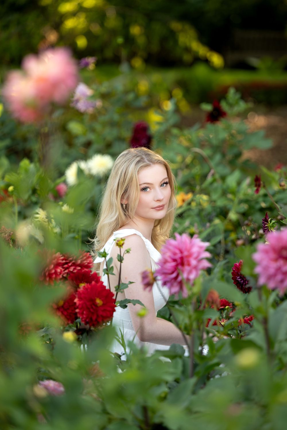 Beautiful blonde woman in white dress sitting among pink and red dahlia flowers in a garden setting.