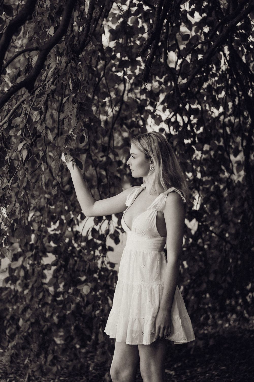 Blonde woman in white dress reaching for branches in a black and white outdoor portrait.