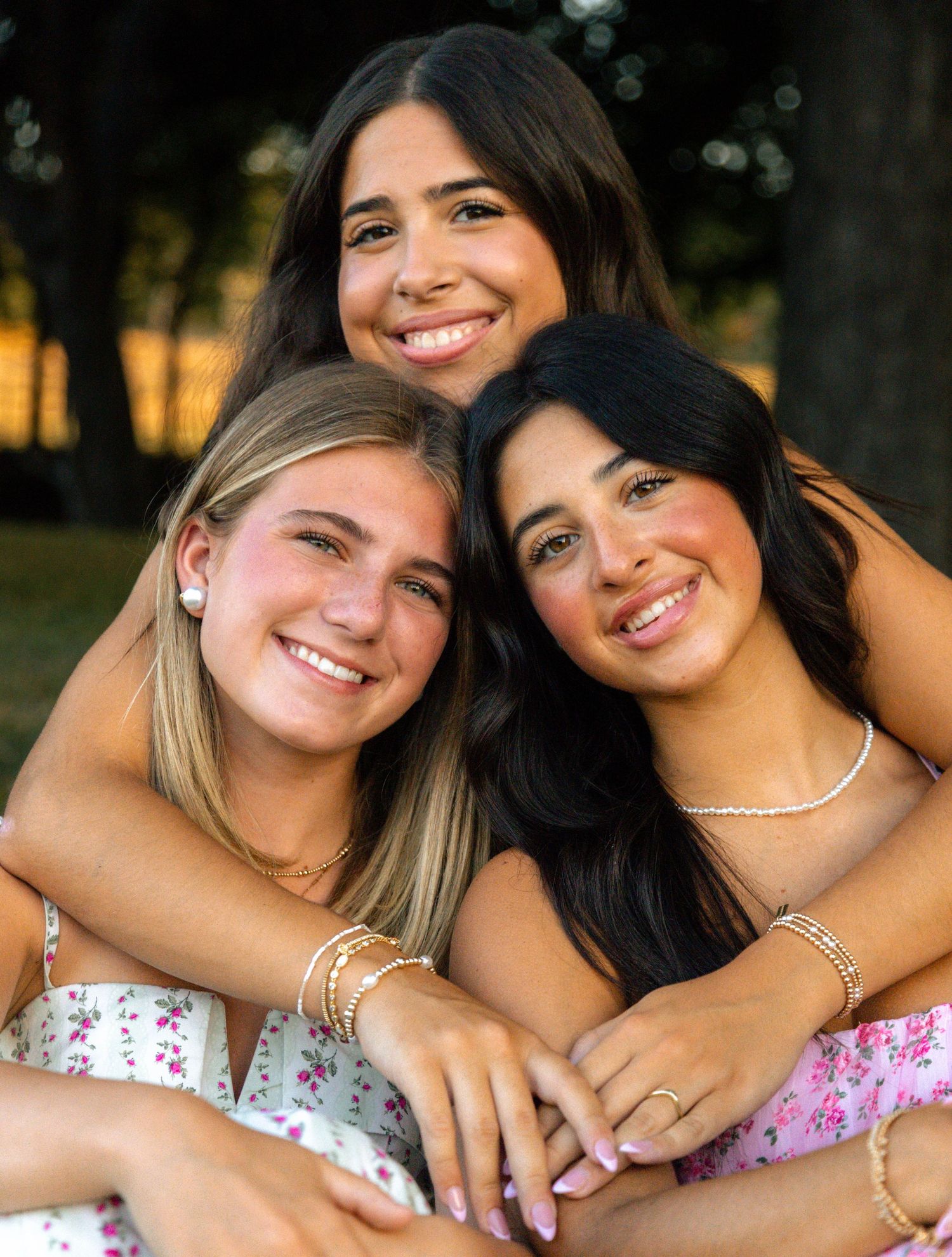 Three smiling young women pose together outdoors in floral outfits during golden hour.