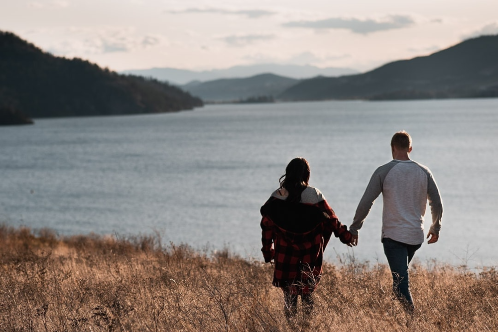 Couple holding hands walking along a scenic lakeside trail with mountain views at sunset.