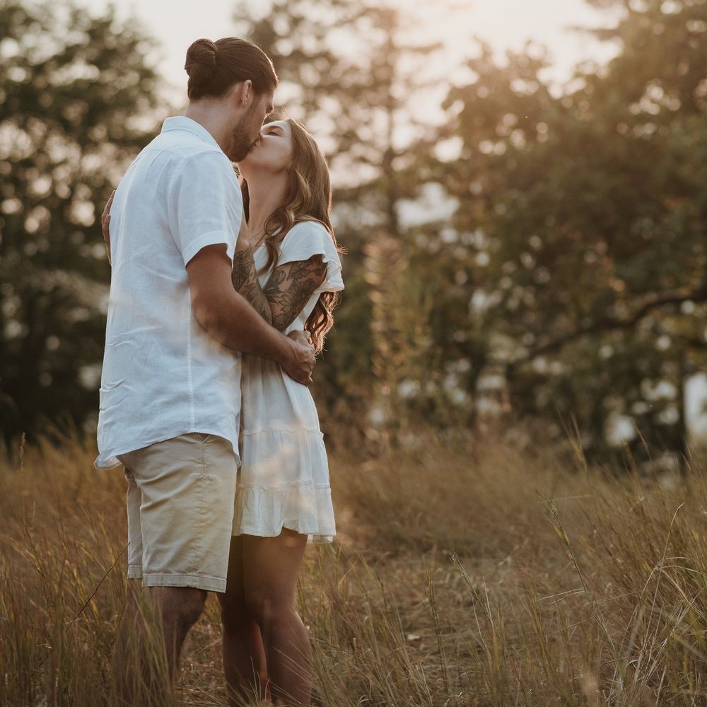 A couple shares a romantic kiss in a golden sunlit field surrounded by tall grass and trees.