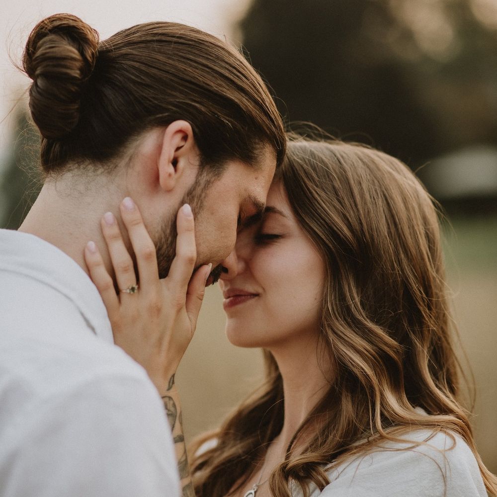 A couple with foreheads touching, sharing an intimate romantic moment outdoors in a soft bokeh natural setting.