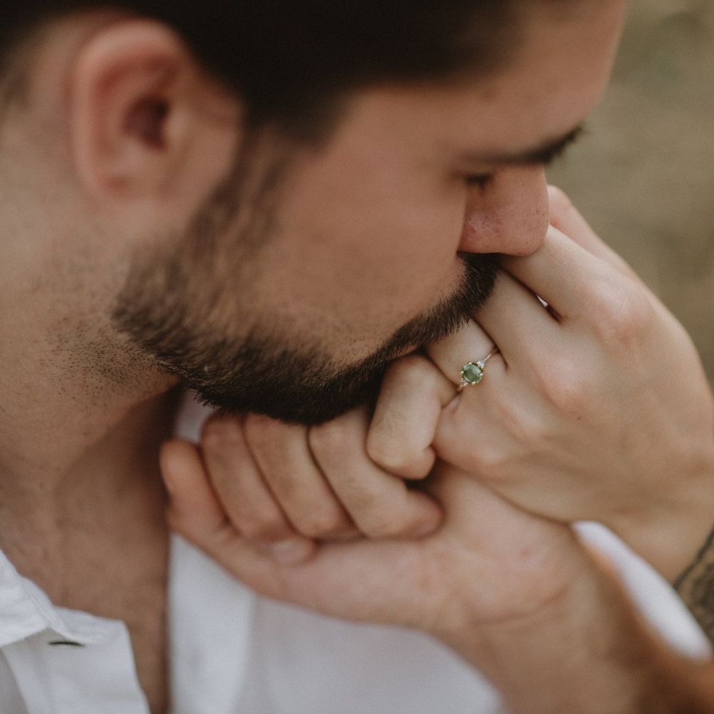 Close-up of a man kissing a woman's hand adorned with a green gemstone engagement ring outdoors.